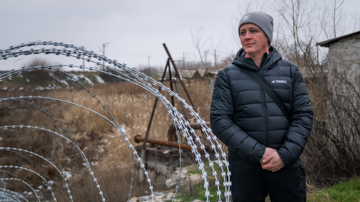 Oleksandr, a man wearing a hat and a dark jacket, stands next to barbed wire.