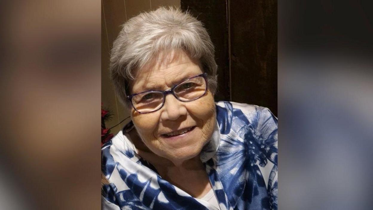 Daphne Hegarty smiling at the camera in a family photo. She has short, grey hair and is wearing glasses with a dark purple frame. She is wearing a blue and white patterned blouse over a white top.