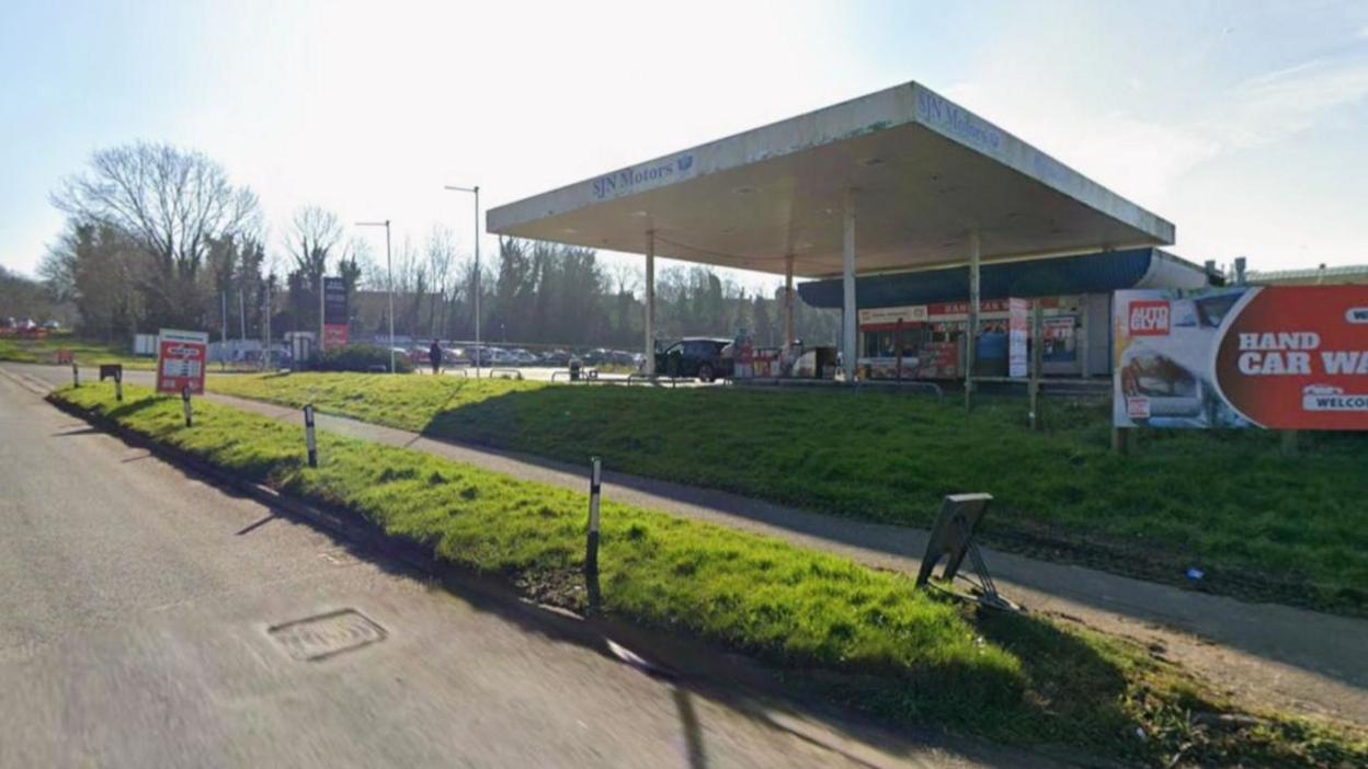 An old fashioned petrol station with a canopy, by the side of a road. There is a grass verge between the road and the petrol station, which says SJN Motors in blue. There is a large freestanding sign for a Hand Car Wash. Next to the petrol station is a car park and trees.