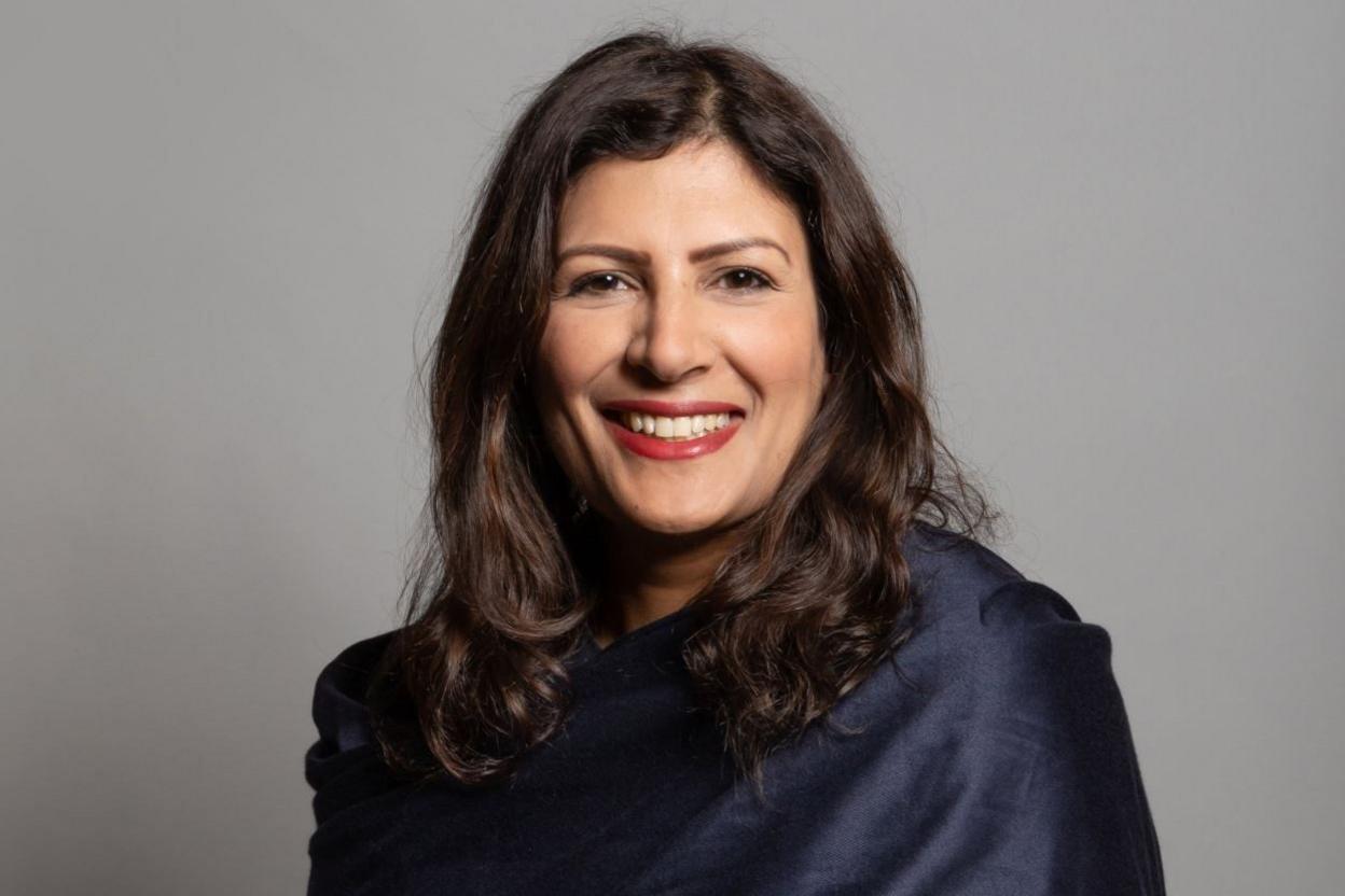 A woman with long dark hair smiles into the camera. She has red lipstick on and is wearing a dark blue top