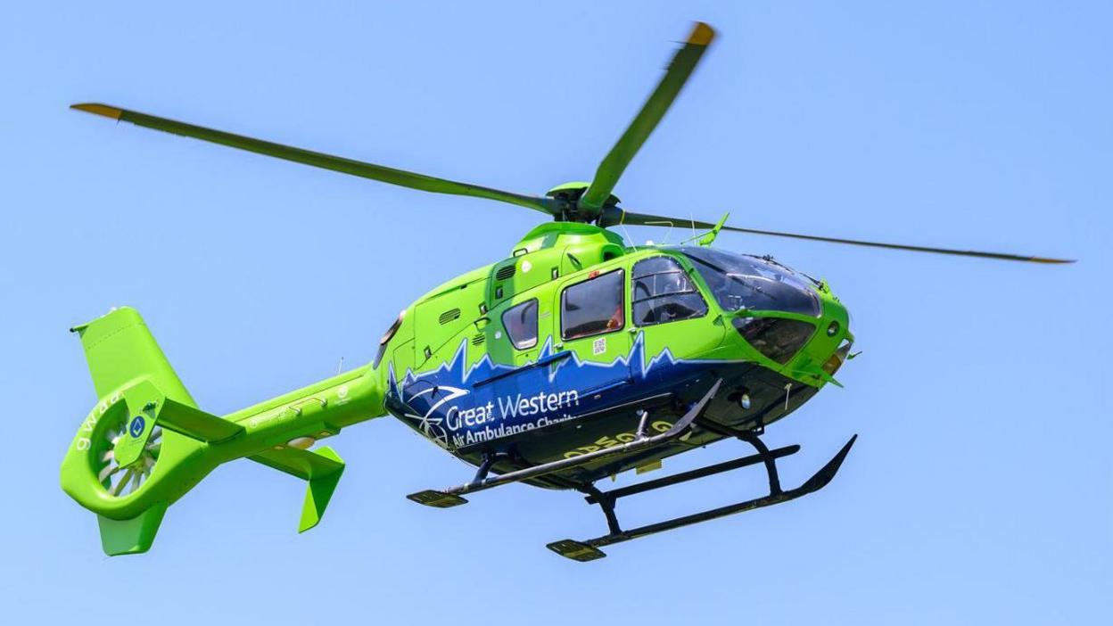 A bright green and blue helicopter hovering in the sky. There is Great Western Air Ambulance Charity branding across the helicopter.