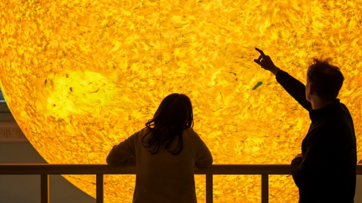 This picture shows two people observing a large, detailed visual representation of the Sun. One person is pointing toward it, while the other leans on a railing, looking at the display. The Sun is illustrated with vivid yellow and orange tones, featuring textures that resemble solar flares or sunspots.