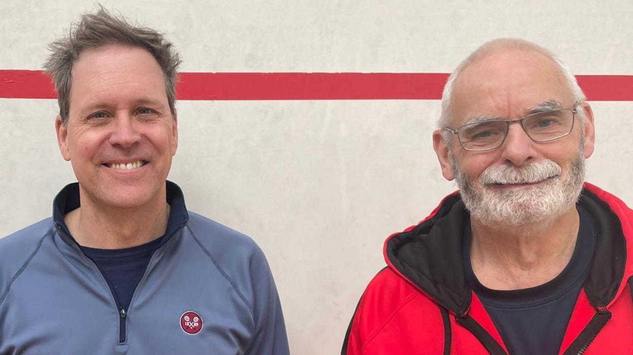 A man in a blue zip top stands next to a man with glasses and a beard in a red zip top both smiling at the camera. 