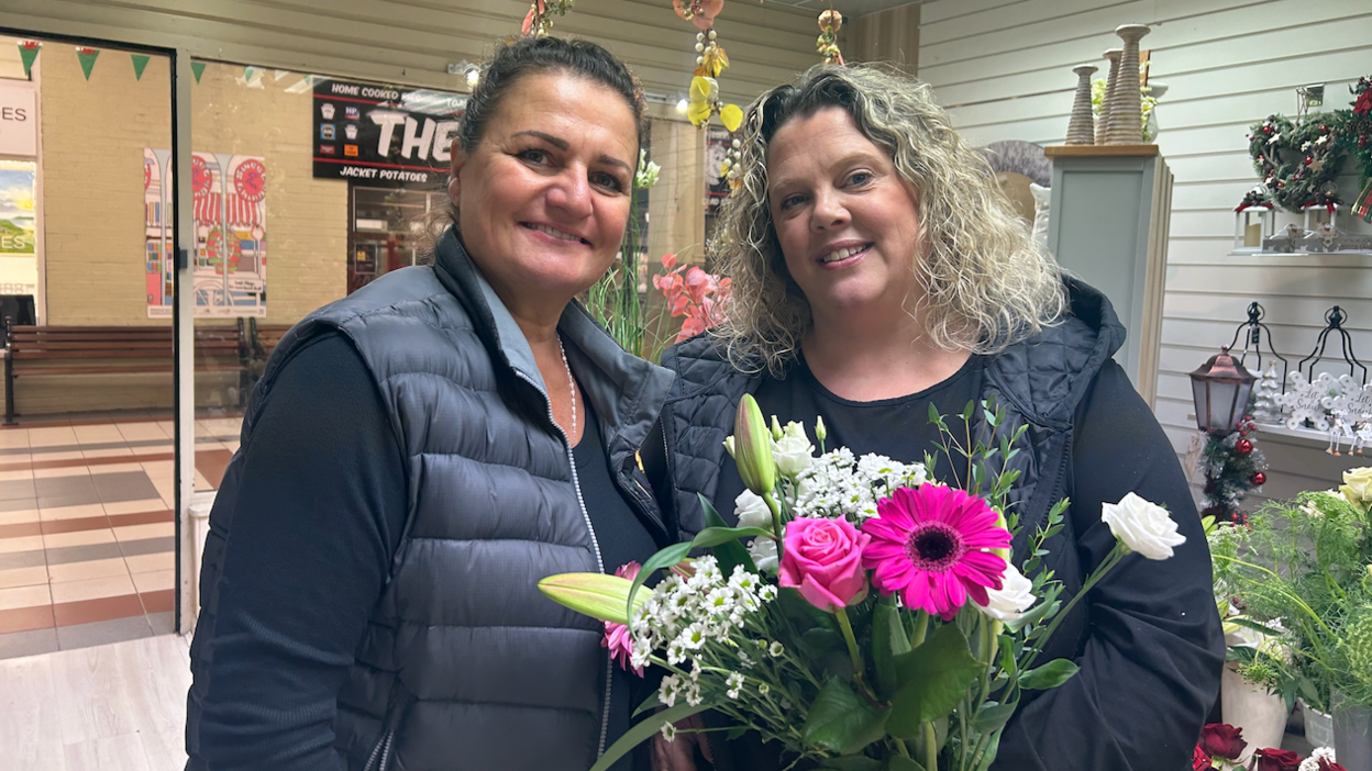 Jane McDonald (left) in a black long-sleeve top and black gillet. Becky Foden (right) also in a black long-sleeve top and black gillet. Becky is holding a bouquet of pink and white flowers, including gerbers, roses and lillies.