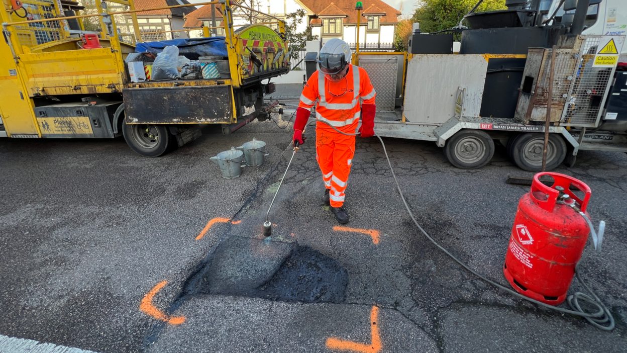 Man in orange high viz with a helmet repairing a pothole. He is using a gas powered heater to work on smoothing the pothole before filling it with material.