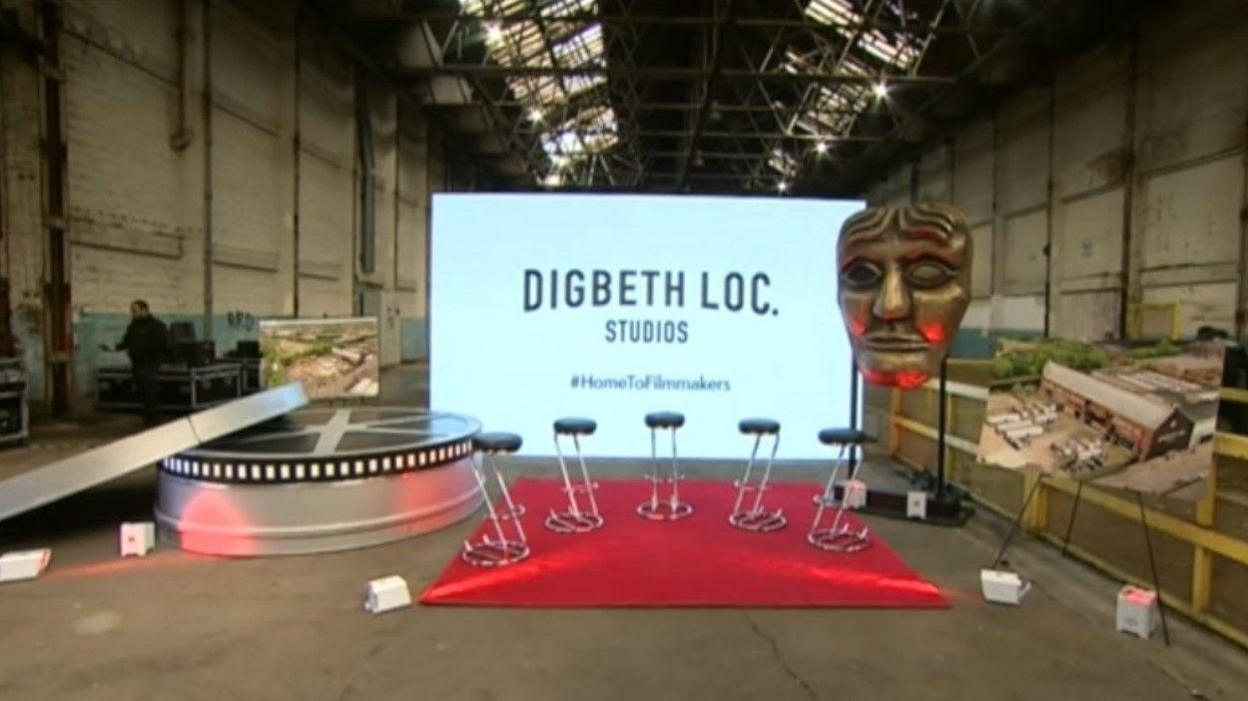 Interior of Digbeth Loc Studios, where there is a movie a screen, a giant film reel canister and an oversized Bafta award behind a series of stools on a red carpet  