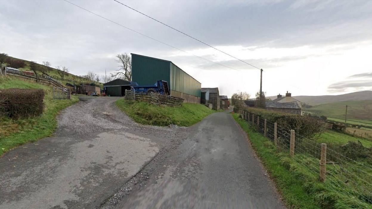 A Google Streetview screenshot of Newlands Farm near Wigton. There is a narrow country road, with a ramp leading to farm buildings on the left. On the right there are fields and open countryside.