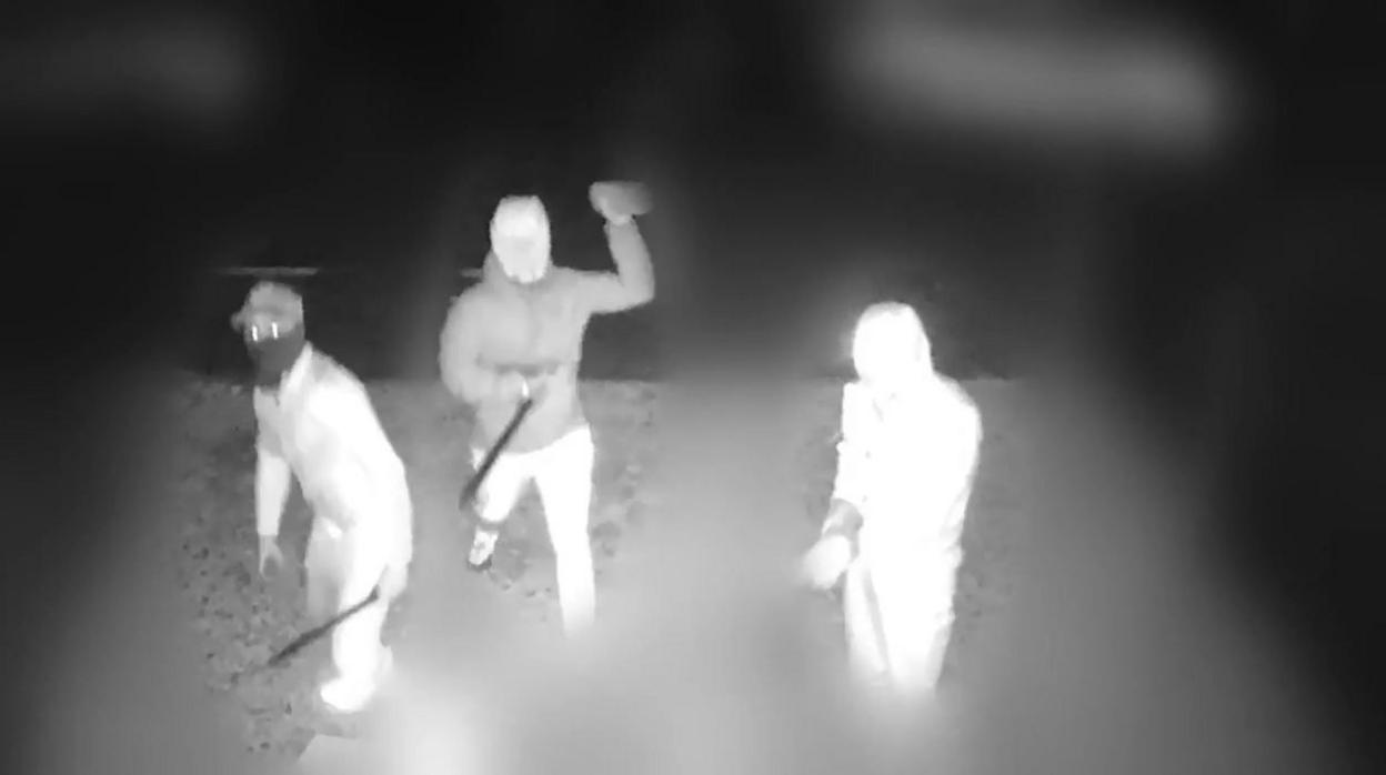 Black and white CCTV still showing three figures, with the rest of the view blurred. The three people, who appear to be wearing face coverings, hats and hoods to obscure their identity, can be seen brandishing machetes as one of them throws a brick.