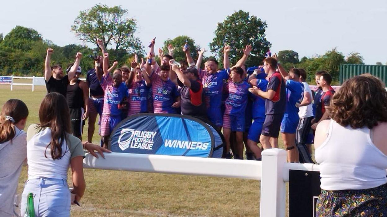 The Anglian Vipers squad and the coaching setup lift the East Rugby League trophy on the pitch at home ground Wymondham Rugby Club, next to an official Rugby Football League stand