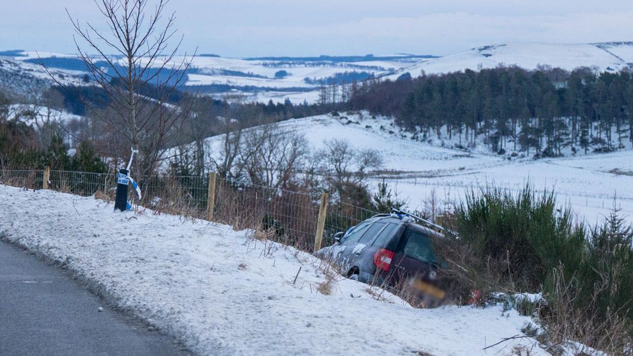 Car in a ditch at the side of the road. The grass verge and surrounding hills are covered in snow. There is police tape wrapped around a tree near the car.