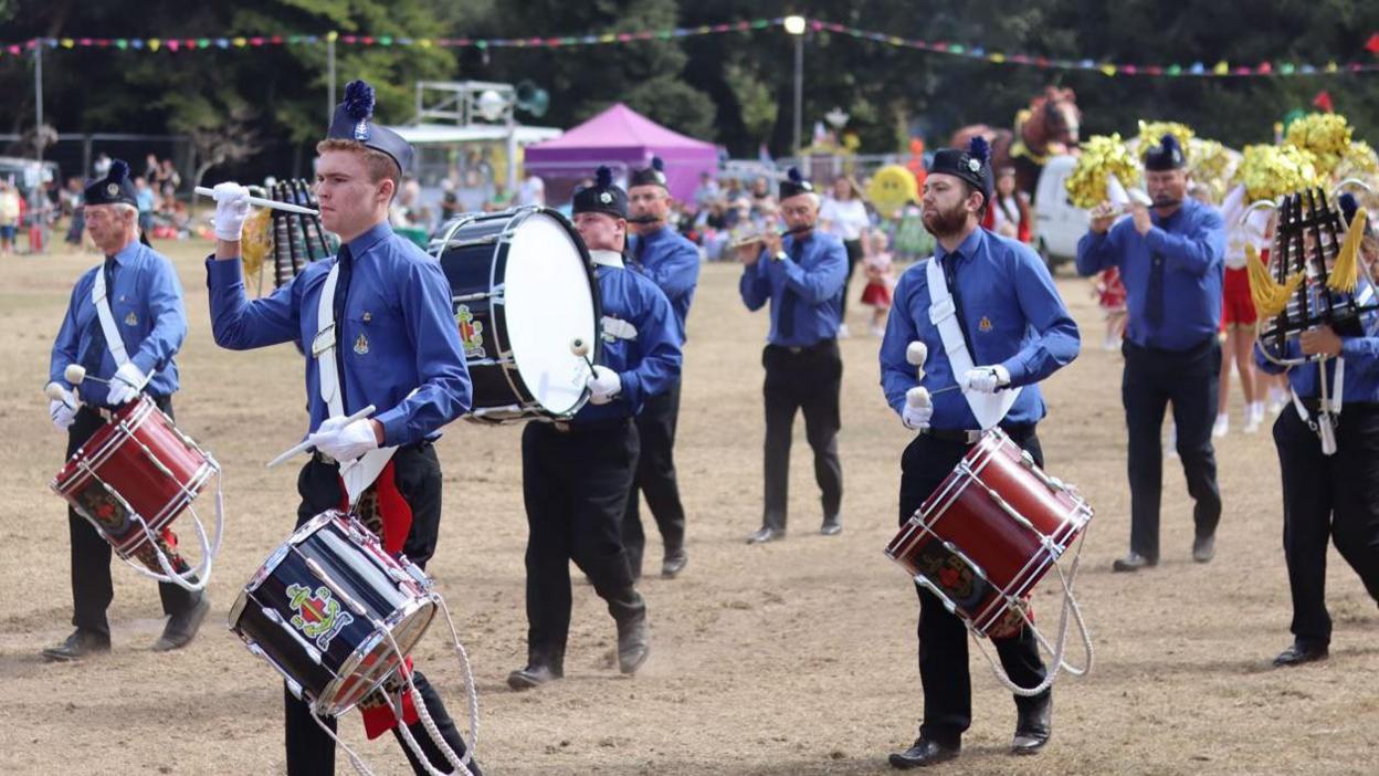 A marching band wearing blue shirts, black trousers and navy blue hats parade at the North Show. Some are carrying drums. Others are playing flutes or wind instruments.