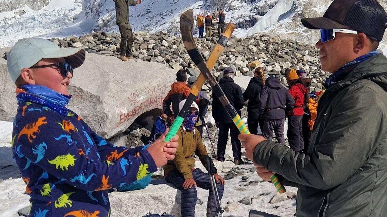 Fergus is wearing sunglasses, a cap, colourful fleece, blue trousers and grey walking boots. He is holding a caman. Opposite him stands a sherpa. who is wearing sunglasses, a cap, green hooded jacket, grey trousers and brown boots. The pair are standing on snow and behind them are high, jagged snow-covered mountains behind him.