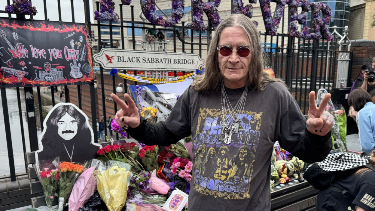 An Ozzy Osbourne look-a-like stands in front of the Black Sabbath bench which is beckeded with bouquets of flowers. He wears a Black Sabbath t-shirt and holds his fingers up in peace signs.