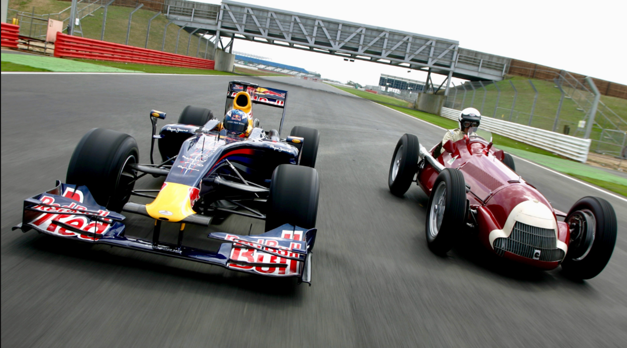 A modern navy blue F1 car drives down a track next to a red 1950s F1 car.