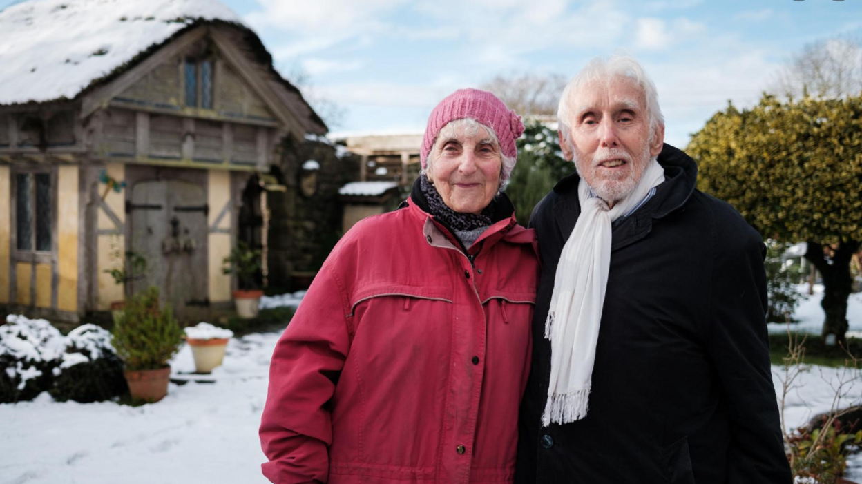 The Buntings standing in the snow in their garden, dressed up warmly, with one of the replica houses, the timber-framed Elizabethan house, in the background