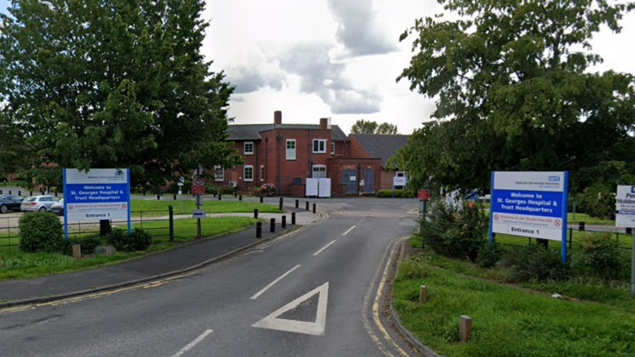 A brick building with a road leading up to it and two blue and white signs either side reading 'Welcome to St George's Hospital and Trust Headquarters' and 'Entrance 1'. Trees can be seen behind the signs and grass all around the road.