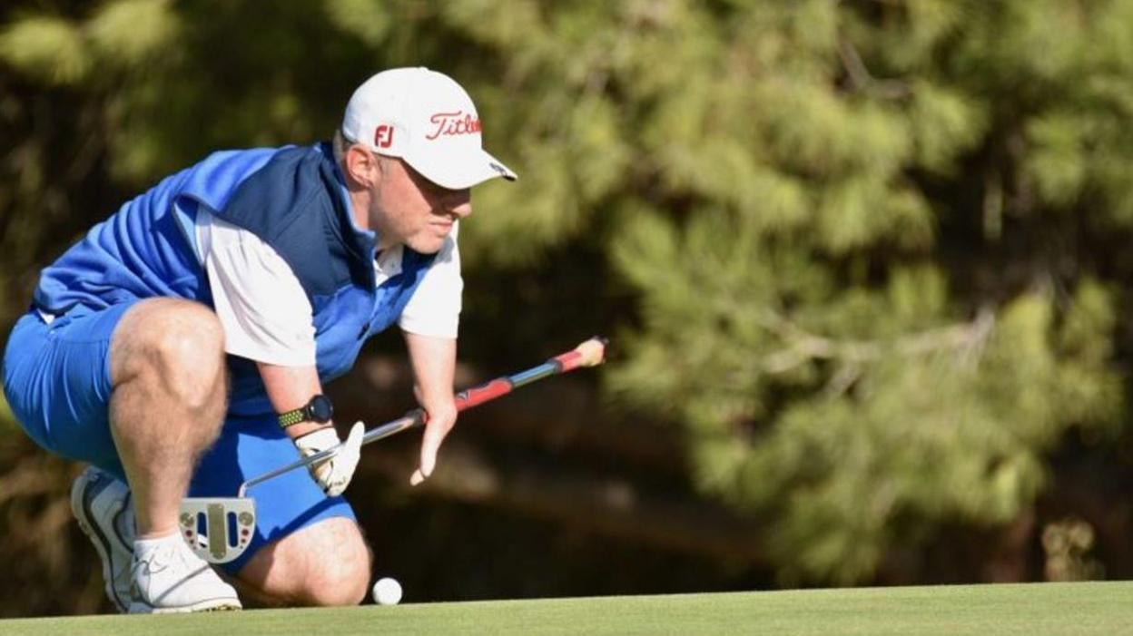A golfer in blue clothing and a white cap crouching down with the ball at his feet.