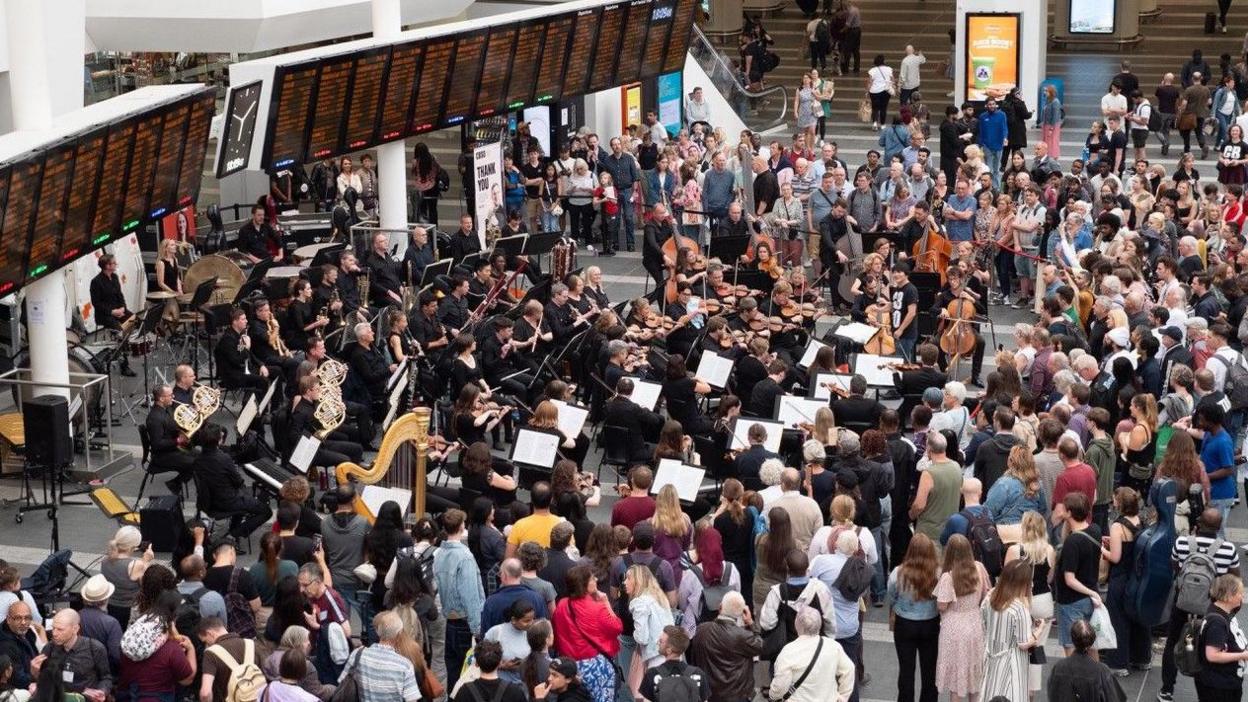 The view down towards the departure boards at a railway station. Members of an orchestra play in front of the signs, with a large number of people surrounding them.