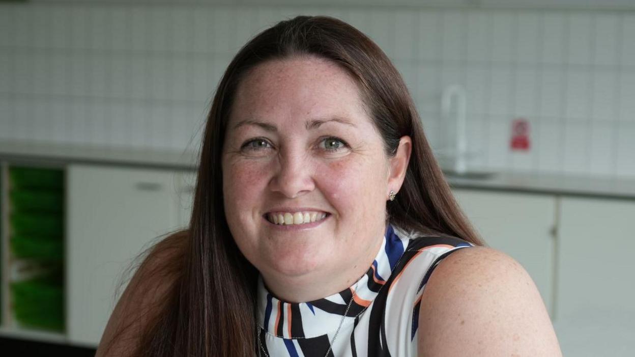 Tracey Plane wearing a sleeveless top, which is white with abstract black, orange and blue stripes. She has long brown hair and is smiling while sitting inside a classroom with white cupboards and a tiled wall.