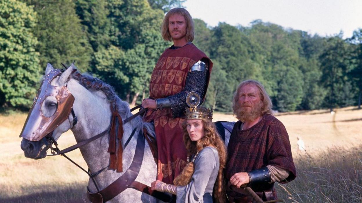 People in period costume - one on a horse and two beside it, one a woman in a crown, standing in a field in the countryside