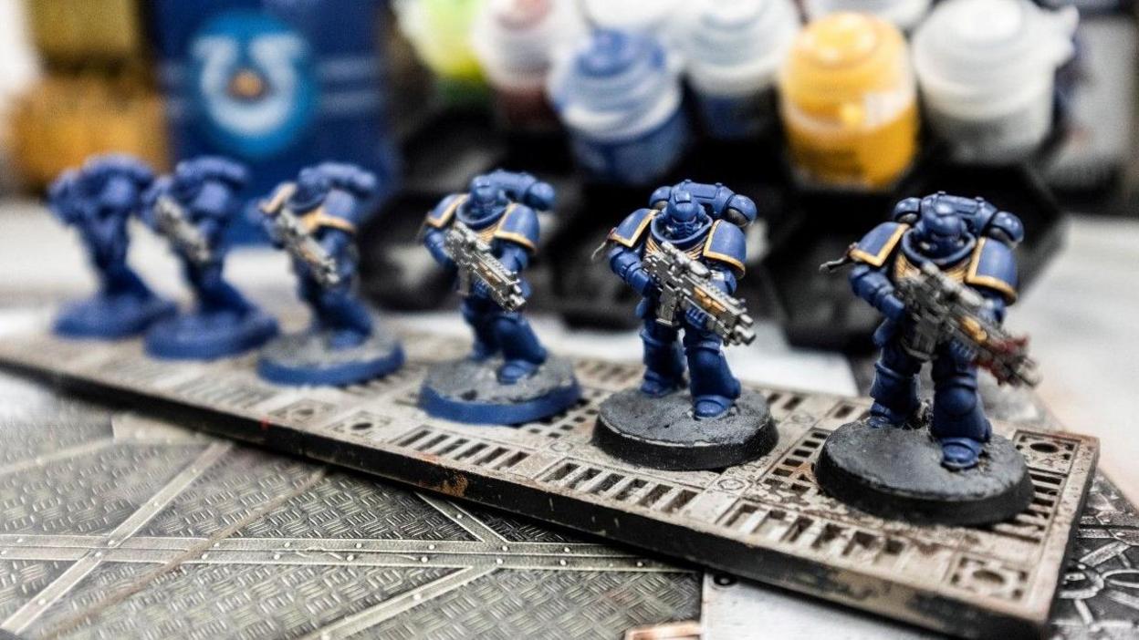 Close up of a series of identical figures, depicting armour clad soldiers, on a table next to some paints