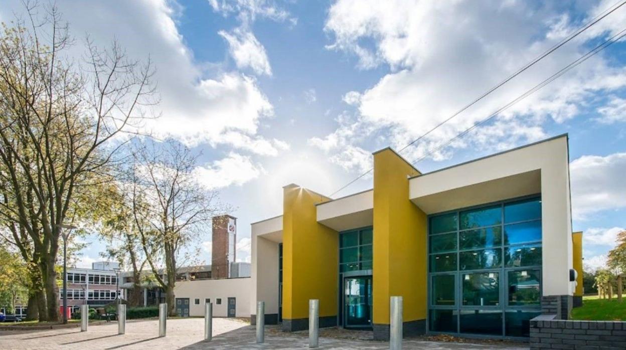 A building with prominent yellow pillars is on the right. In the distance is a brick tower and on the left there is a three-storey building in the background behind trees.