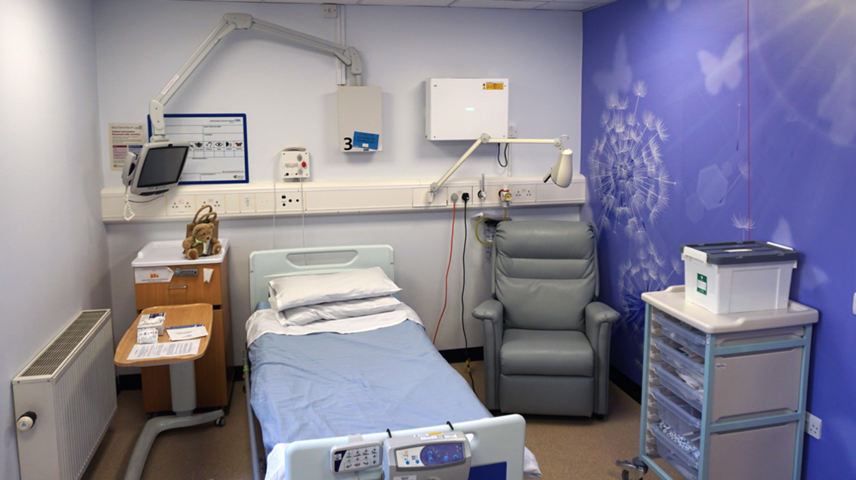 A hospital room showing a white single bed with a blue blanket, with a basin on its left and a blue padded chair on its right, with a clinical set of drawers just ahead. On the white wall behind it is a monitor screen on an arm and a lamp on an arm. The wall on the right is blue with white dandelion clocks and butterflies.