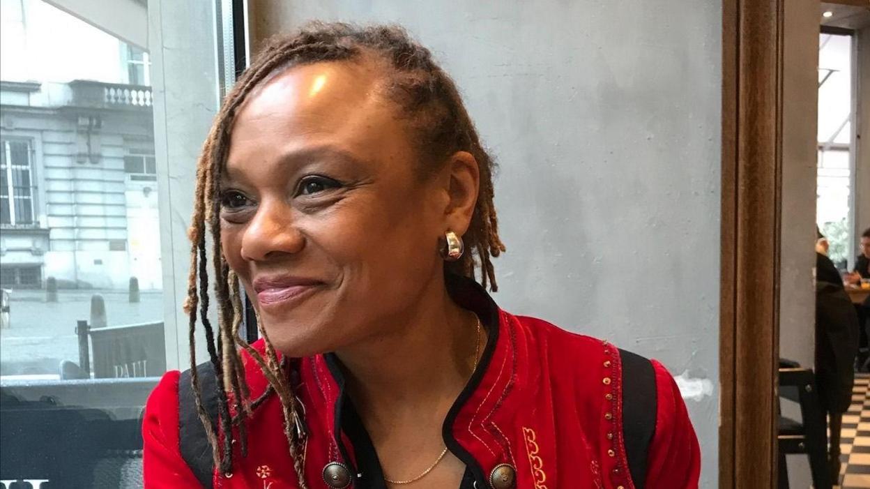 Dr Laura Fish, smiling, wearing a bright red jacket and sitting in a coffee shop. She has gold earrings and her hair is tied into lots of thin braids.