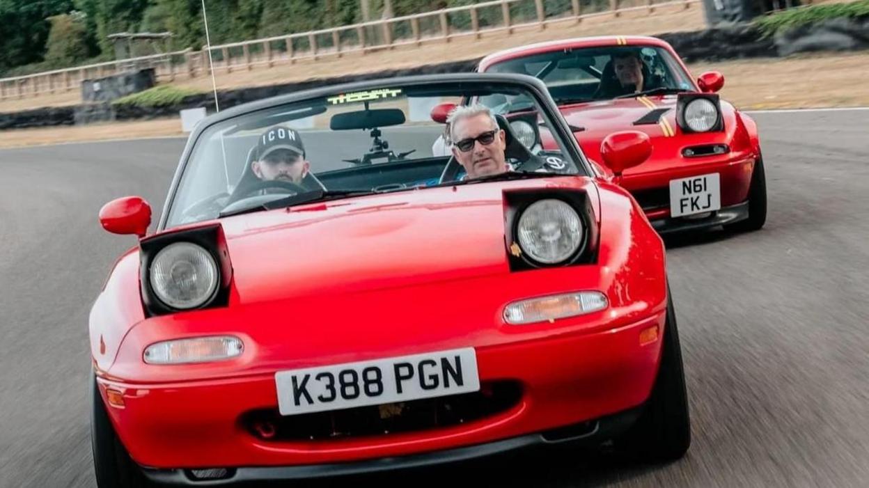 Two red Mazda sports cars with pop-up headlights on a race track. Two people can be seen in the first car, which has its roof down.