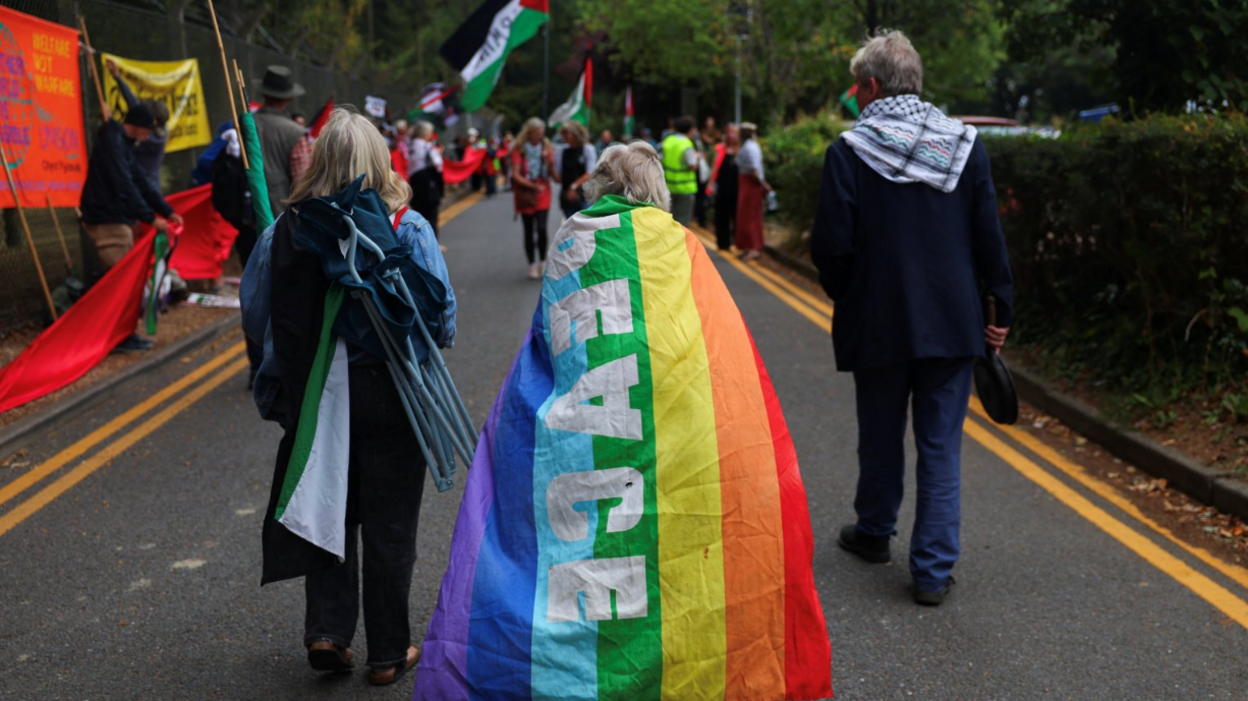 Three people walking away from the camera along a single-track road. A woman carries a camping chair on her back, another woman wears a rainbow flag as a cape with "PEACE" in white letters on it, and a man is wearing a black jacket and jeans. All three have grey-blonde hair. To the left is a series of flags along a fence carrying unreadable slogans, with people in the background carrying flags.