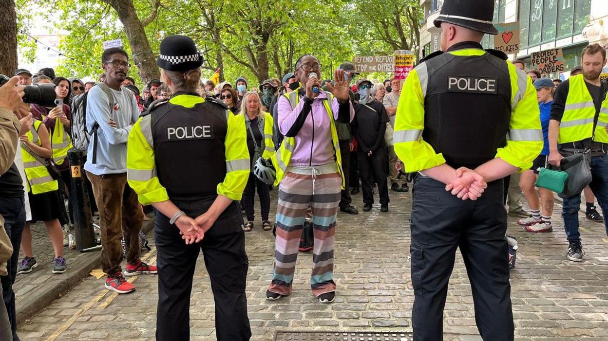 Police with their backs to the camera holding a line in front of a large number of counter-protesters. One woman is shouting into a megaphone in front of them.