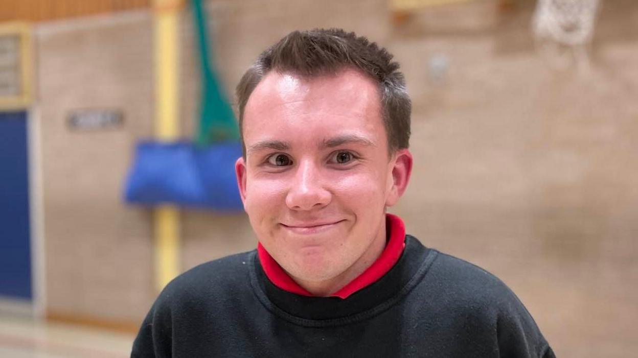 Hamish Crawford smiles at the camera while standing in a sportshall. He has short brown hair and is wearing a black jumper with a red polo top on underneath.