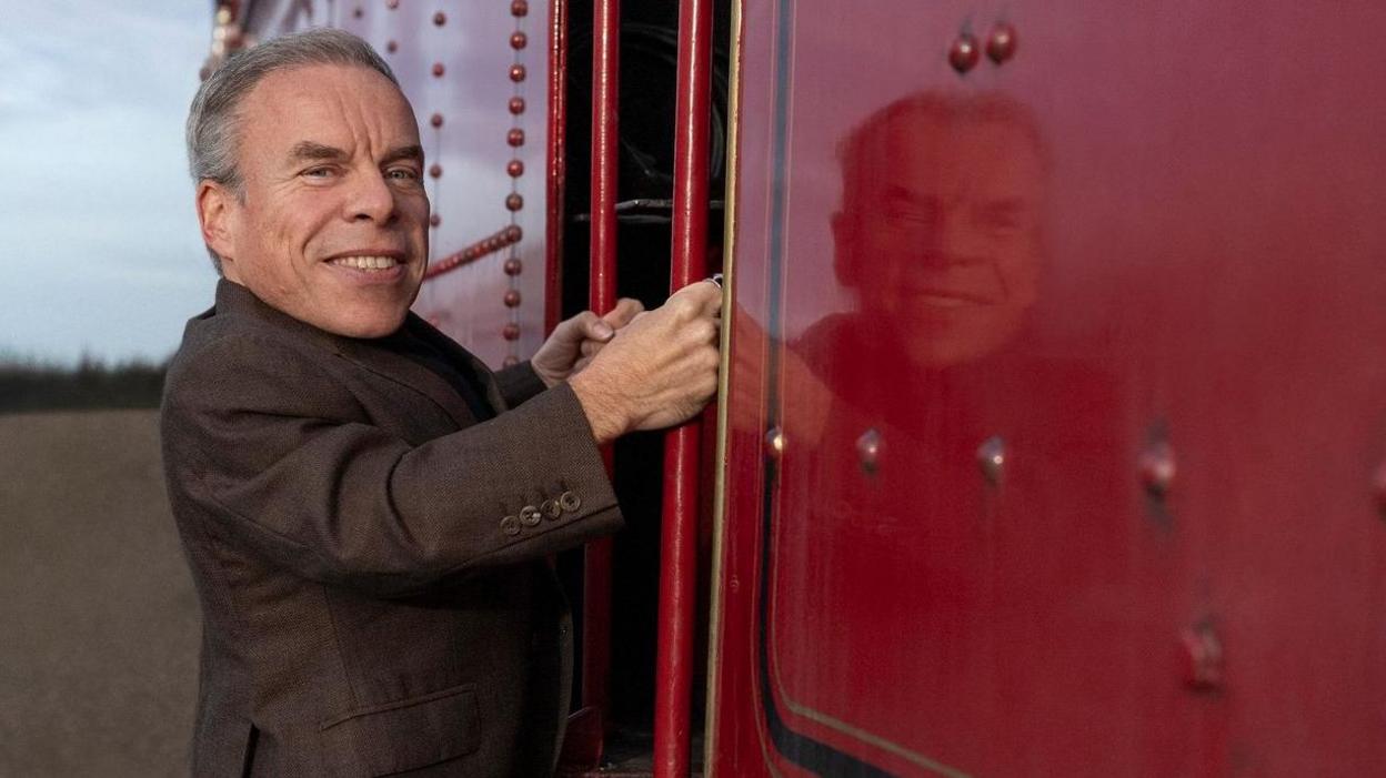 Warwick Davis boarding a red steam train and smiling at the camera