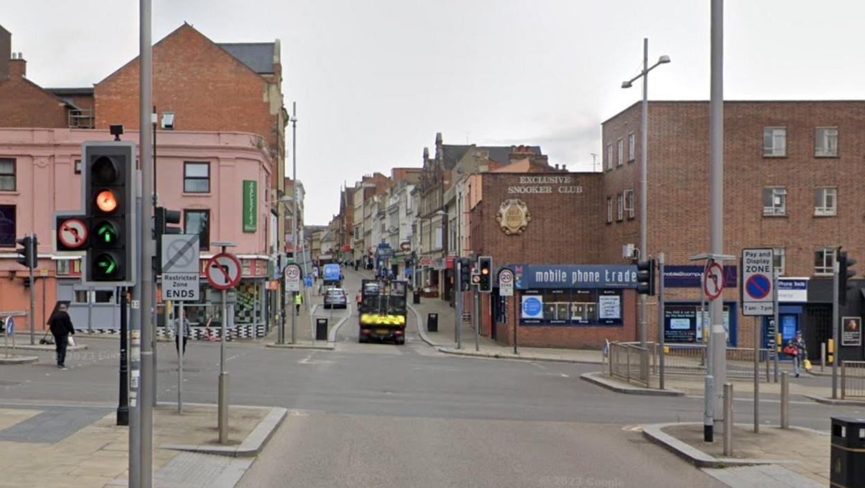 Road junction with traffic lights and "no left turn" signs. A lorry is visible up ahead, and there are shops on streets around the junction, including one selling mobile phones.  A person is walking across the road to the left.