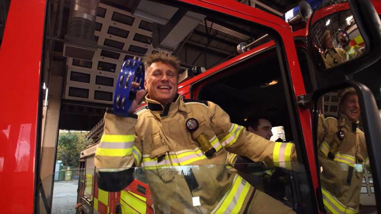 A firefighter leans out an engine and shakes a tambourine.