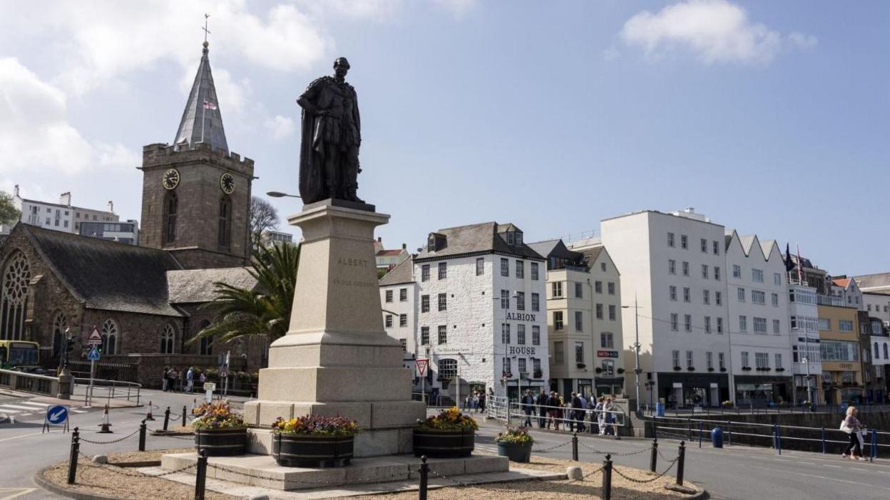 People walking around Albert Pier and Victoria marina at St Peter Port on a sunny day with the Albert statue prominent in the skyline in front of a church.