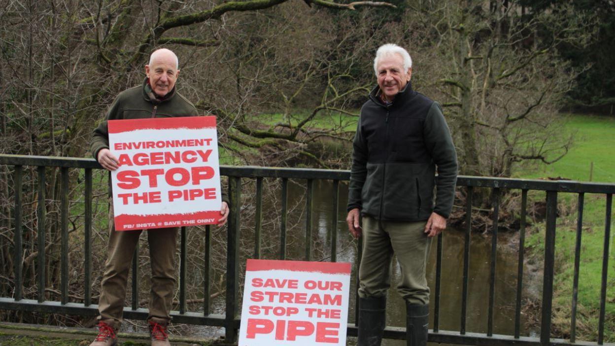 Two men with thinning grey hair standing in front of railings with red and white signs which read save our stream, stop the pipe