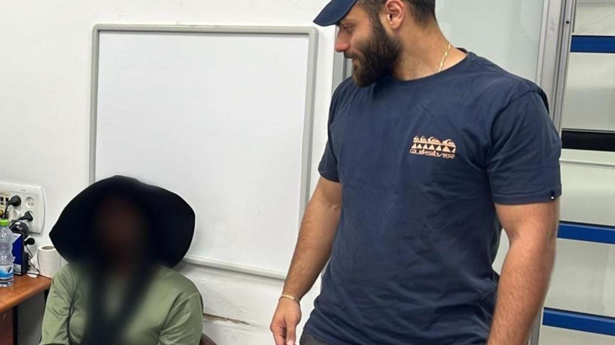 The face of a woman seated wearing a green shirt is blurred. Next to her stands a man in a blue t-shirt and cap.