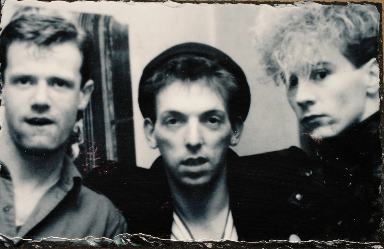 A black and white photograph shows the heads and shoulders of three men looking at the camera. Frankie in the middle has a hat on while Georgie is wearing an open-collar shirt.