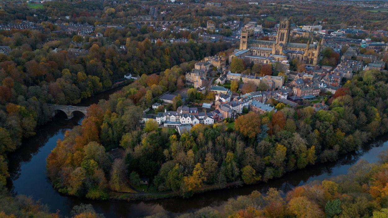 A view above Durham City. It shows Durham Cathedral, surrounded by houses and autumn leaves on the trees along the banks of the River Wear.