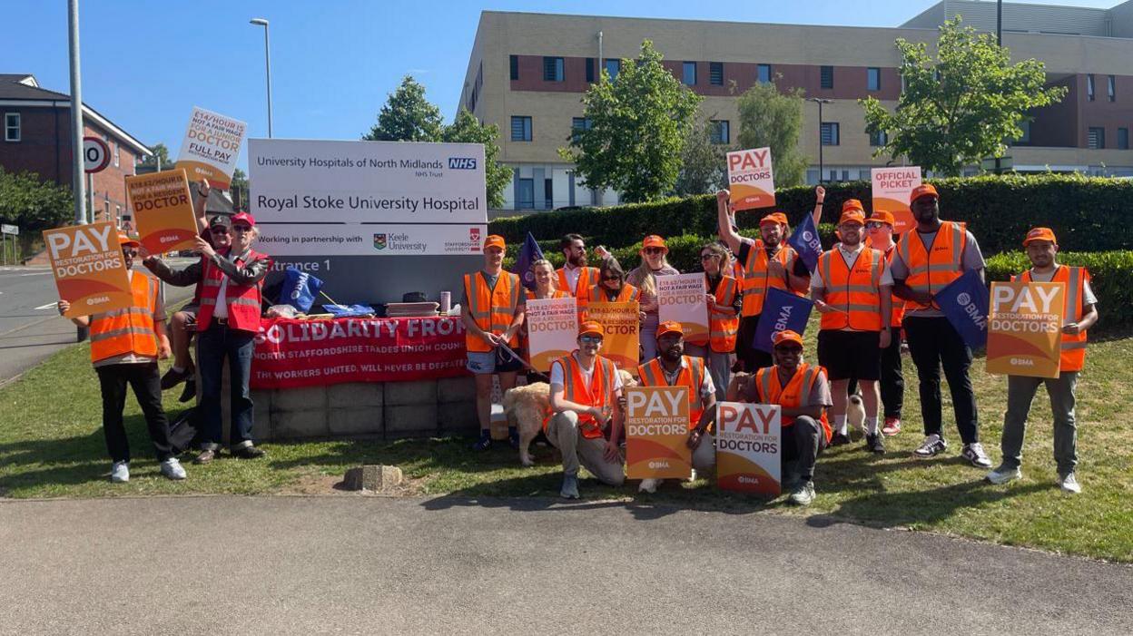 Dozens of people stand outside a hospital building holding placards and signs that say 'Pay Doctors' and wearing orange hats. A large white sign with the words 'Royal Stoke University Hospital' is behind them.