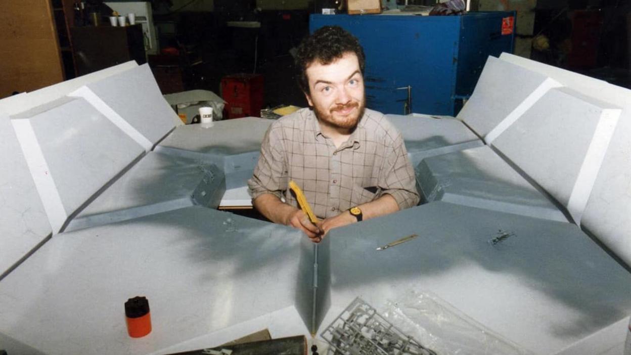 A younger Mike Tucker, with dark hair and beard, is smiling from the middle of a large spaceship model he is building.