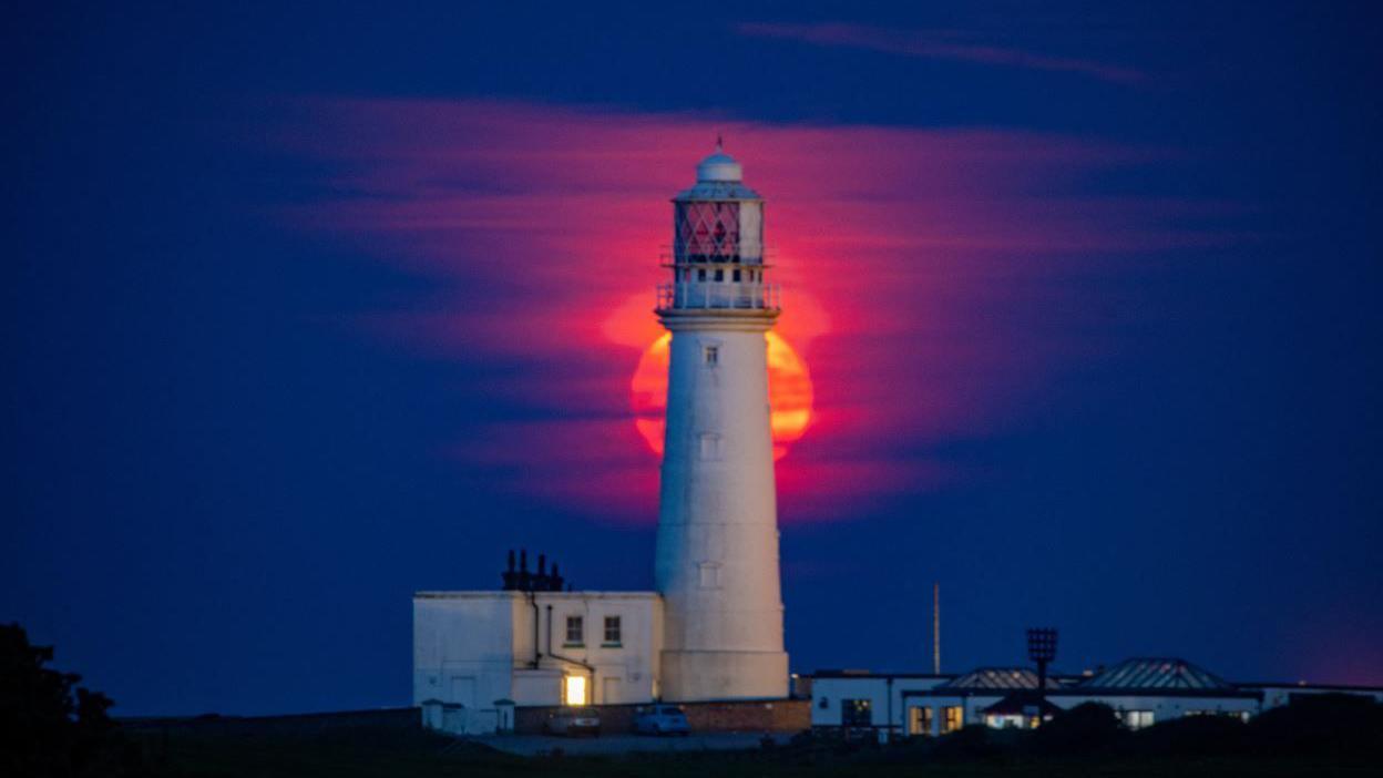 A red-coloured moon behind a white lighthouse. It is night time and the sky is a dark blue. 