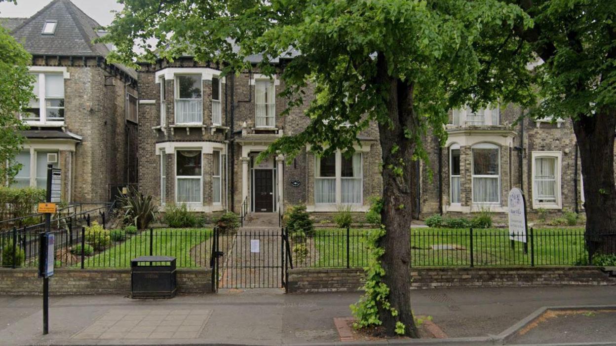 A two-storey grey brick building with a dark-coloured front door and bay windows on either side. There is a small black-fenced garden with green grass in front of the building. There is a path with a large tree in front of the garden.