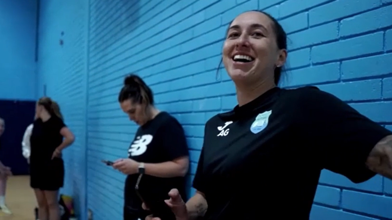 Amie Gray wearing a black sports kit and smiling. She is standing against a blue brick wall.