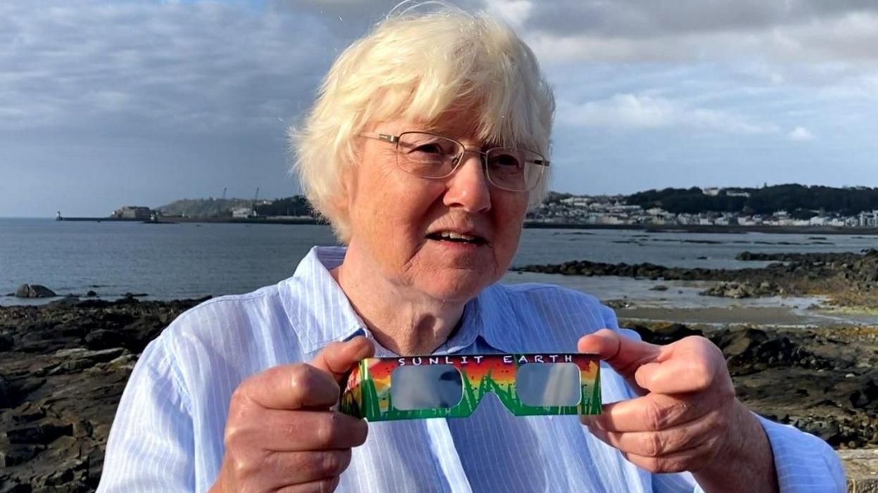 Dr Jean Dean holding a pair of the brightly coloured solar eclipse glasses. She has short grey/blonde hair and is wearing a blue shirt. She also has slim framed silver glasses.
She is stood in front of a coastal view of Guernsey.