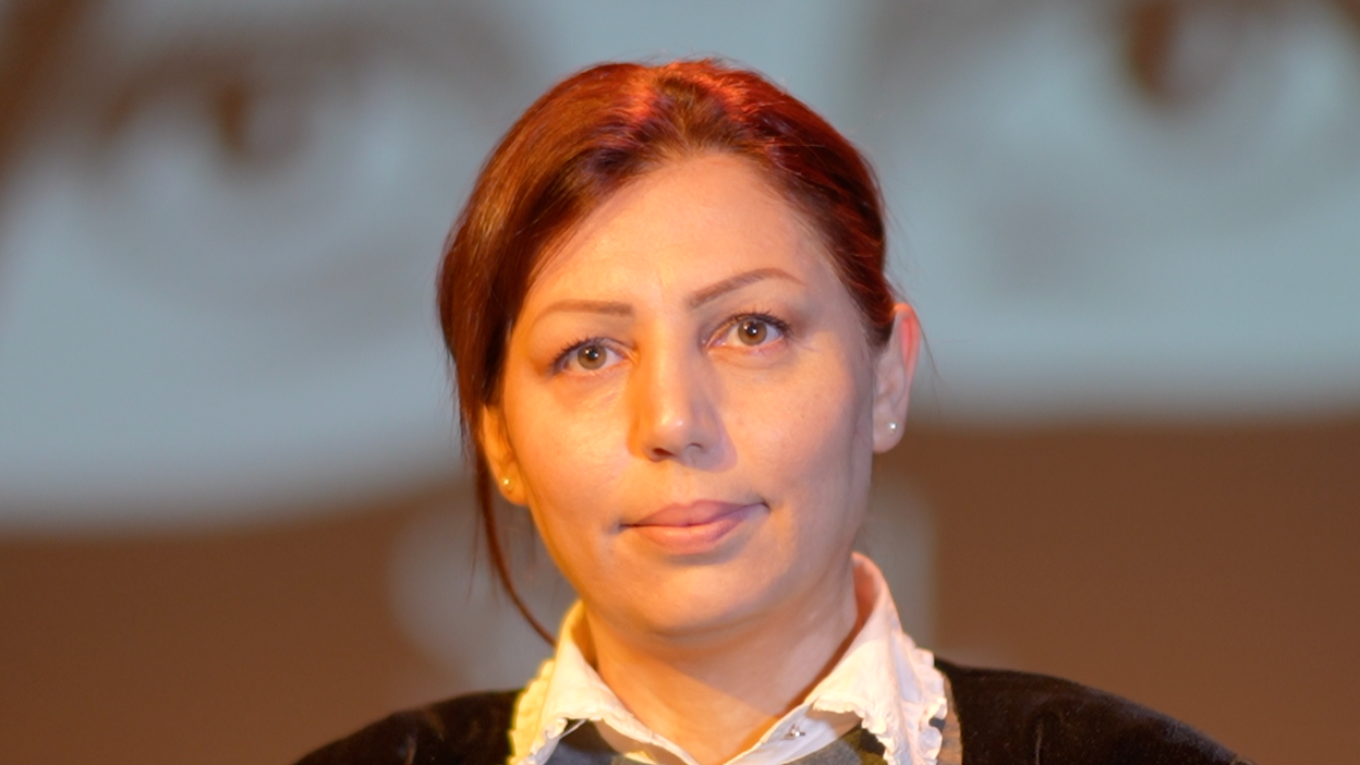 Rona smiles at the camera. She has red hair that has been tied back behind her head. She wears a black jumper with a white top underneath with a collar.