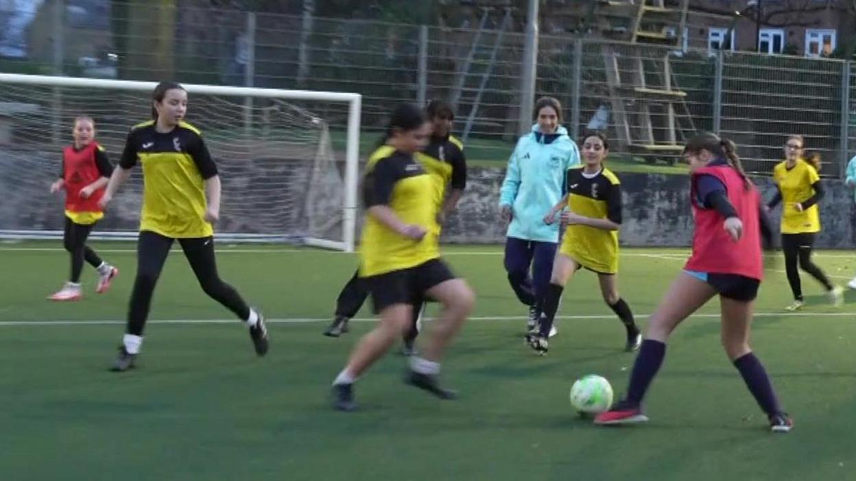 TV screenshot from the edge of a football pitch on which a training session is taking place. Female players wearing red bibs or yellow football tops are kicking the ball.