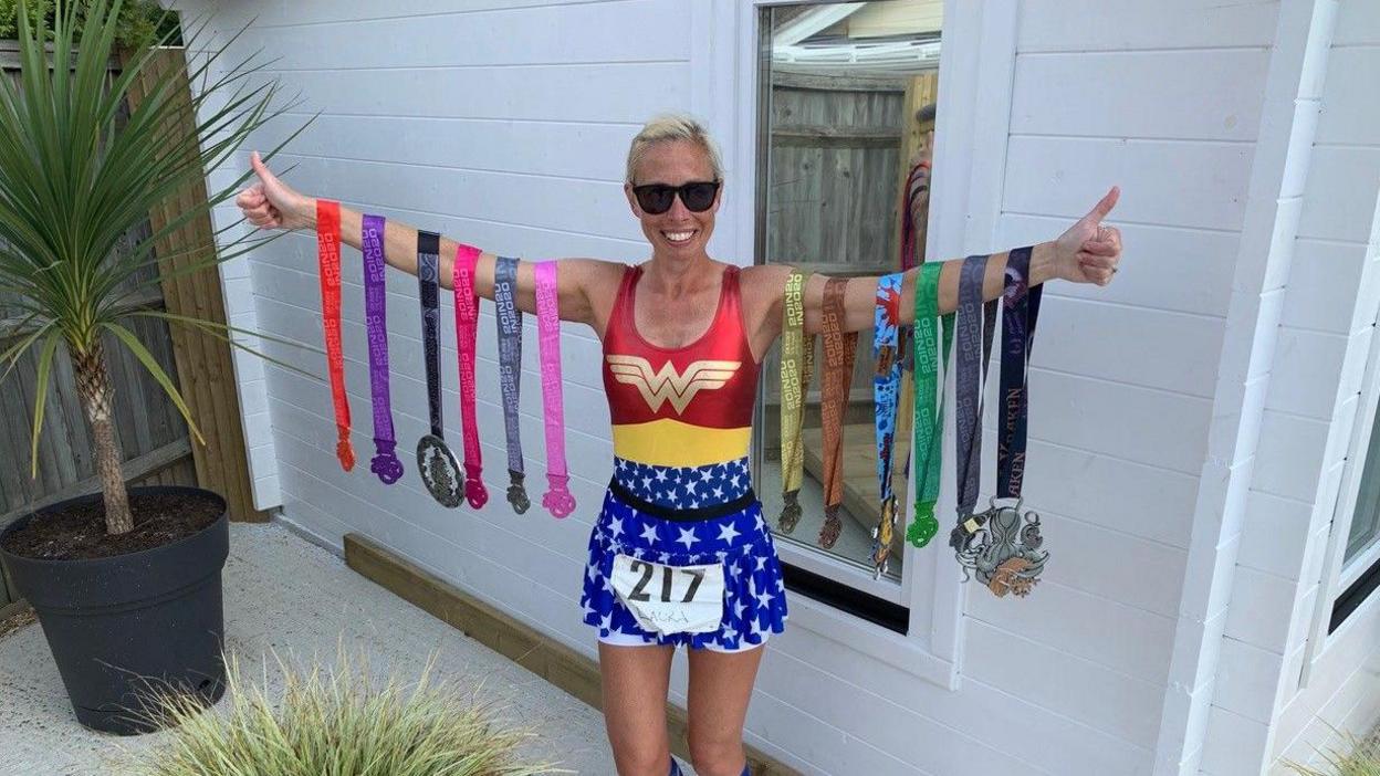 A woman, Laura Watts, has blonde hair that is tie back and her arms are outstretched. On each arm are six medals, all with different colour straps. She is wearing a Wonder Woman outfit of blue shorts and red and yellow top. She has on black sunglasses. She is standing in front of a white wall with a window in it. To her right is a green palm tree in a large black pot.