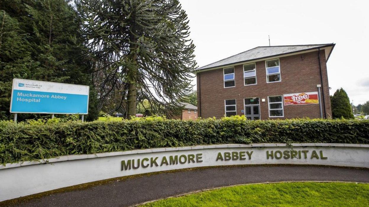 The exterior of Muckamore Abbey Hospital. There is a sign featuring the name of the hospital and it is also written on a low concrete wall. There is a hedge above this wall and a patch of grass in front. Behind it you can see two red brick buildings.